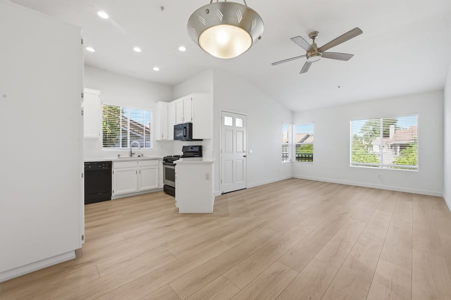 An empty living room with a vaulted ceiling and pale wood floors, opening onto a white kitchen